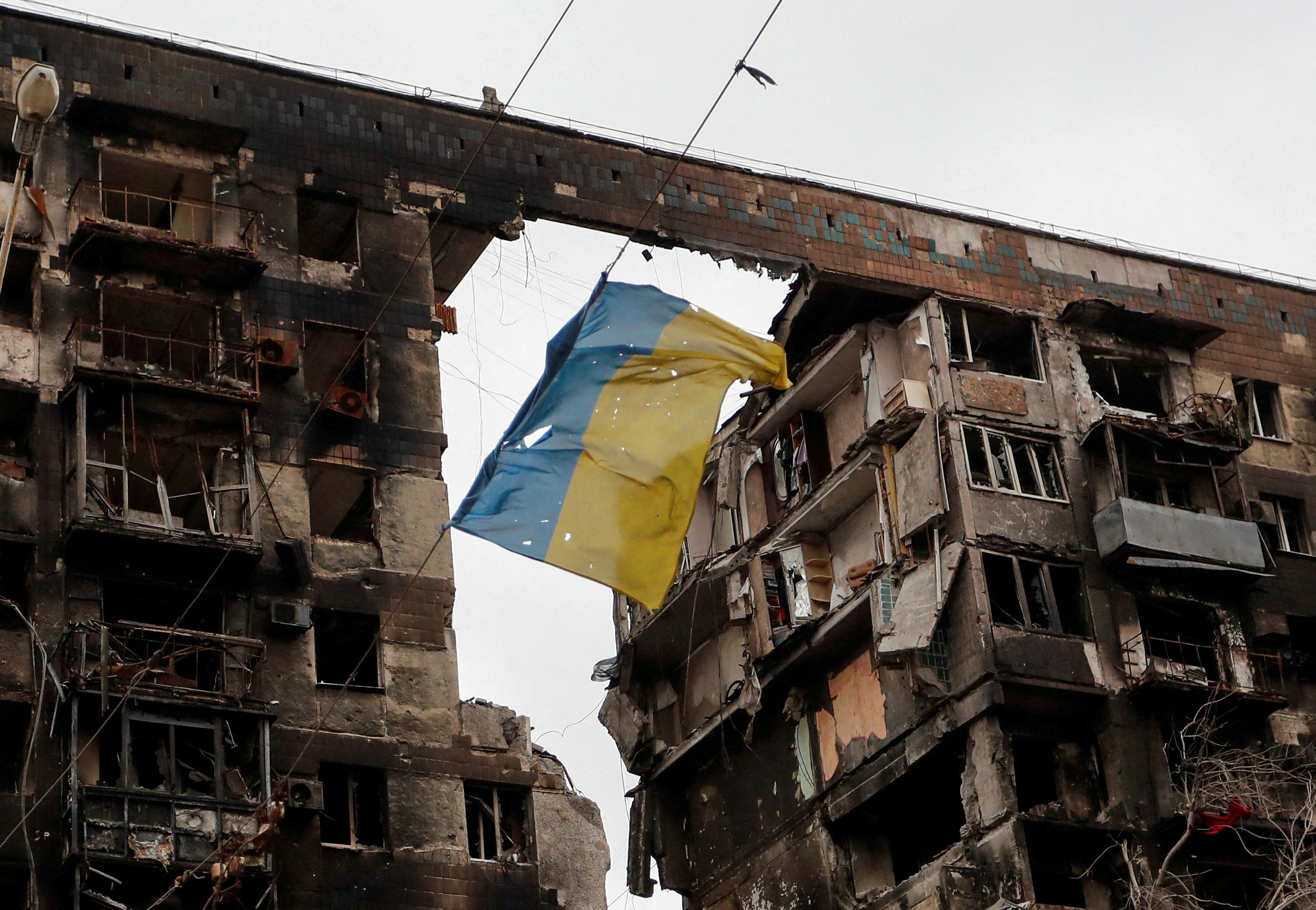 A tattered Ukrainian flag hanging in front of a damaged multi-storey apartment building.