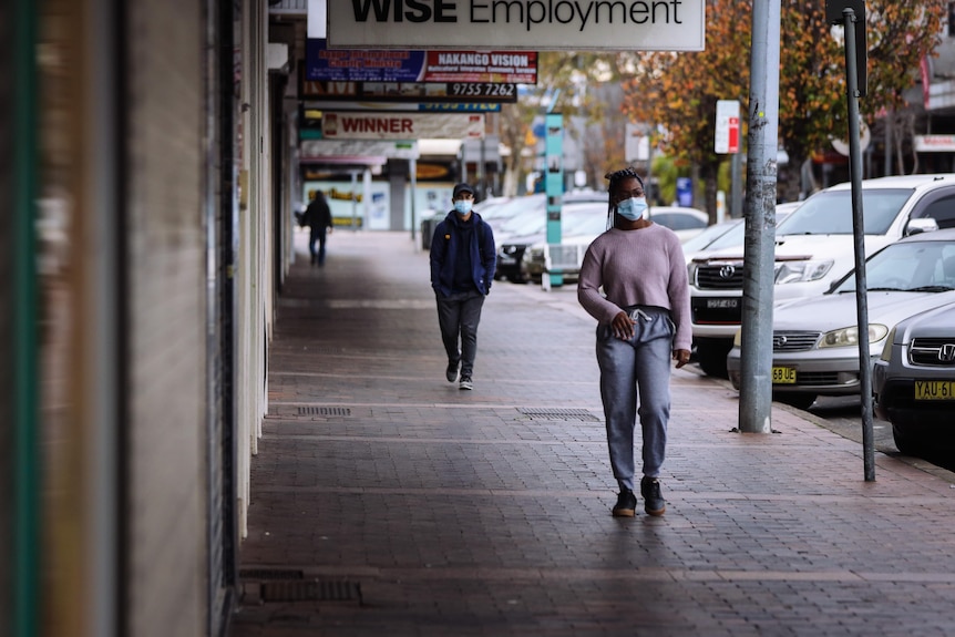 A couple of pedestrians wearing face masks walk past closed shops. 