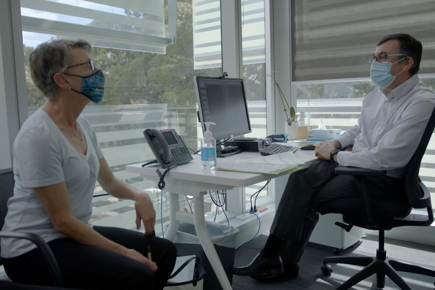 A male doctor and a female patient, both in masks, sit in a clinical room.
