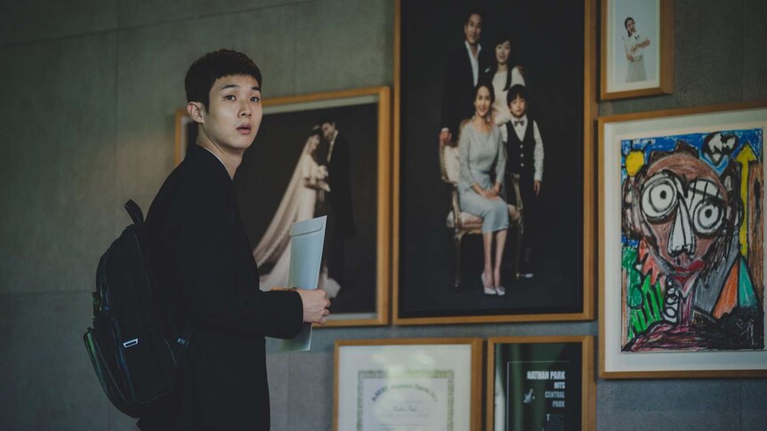 Young man with neat short hair in dark suit stands in front of concrete wall with family photos and children's drawings.