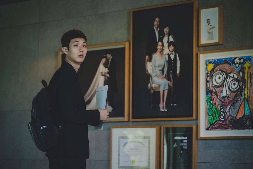 Young man with neat short hair in dark suit stands in front of concrete wall with family photos and children's drawings.