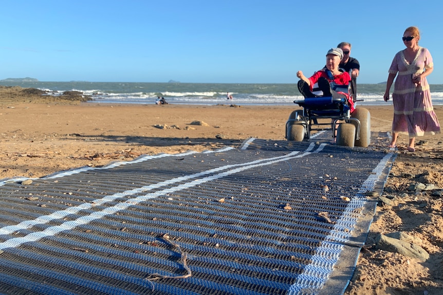 A long mat stretches towards the water on the sand, Mila in the beach chair being pushed, ocean behind.