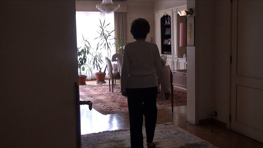 An old woman stands at the threshold of her living room, with her back to the camera.