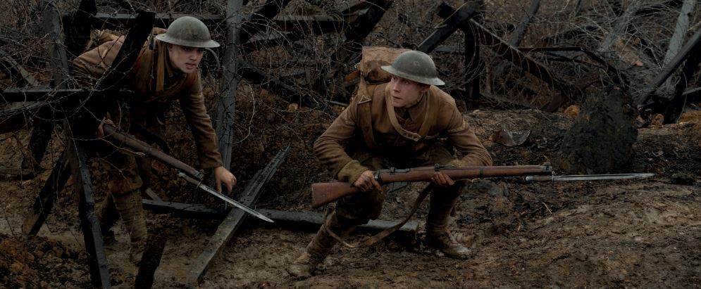 Two men in world war one brown uniform with rifles crouch on a muddy battlefield with wooden obstacles wrapped in barbed wire.