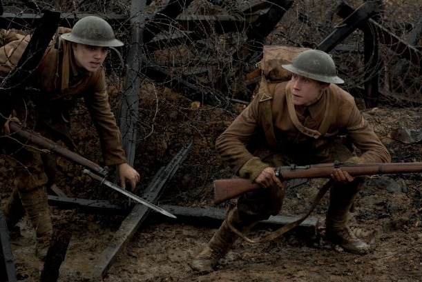 Two men in world war one brown uniform with rifles crouch on a muddy battlefield with wooden obstacles wrapped in barbed wire.