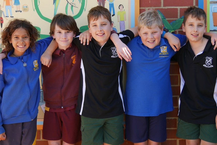 One girl and five four boys from different schools smile in front of a colourful mural with their arms around each other. 