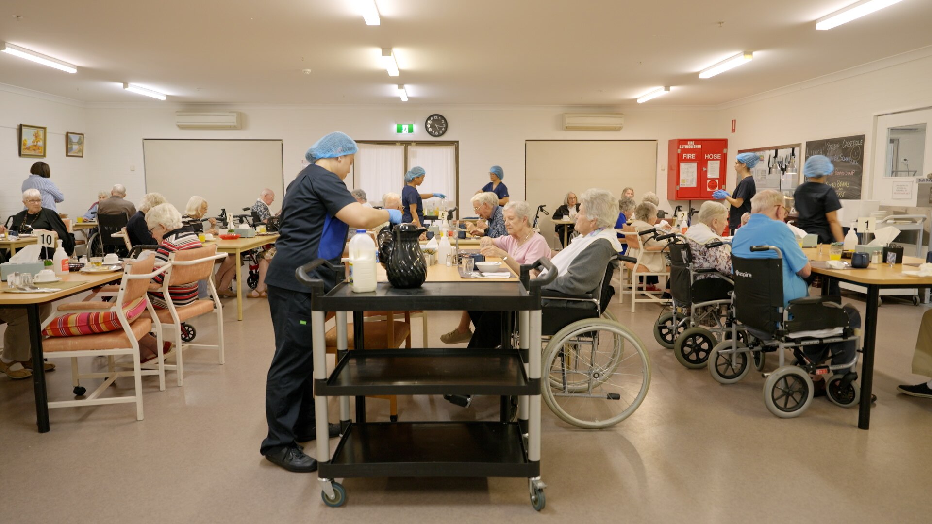 A dining room at an aged care facility with workers in blue hair nets helping serve food to residents