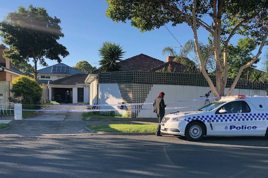 Police tape surrounds a large residential fence in front of a house, as a woman in a suit stands by a police car.