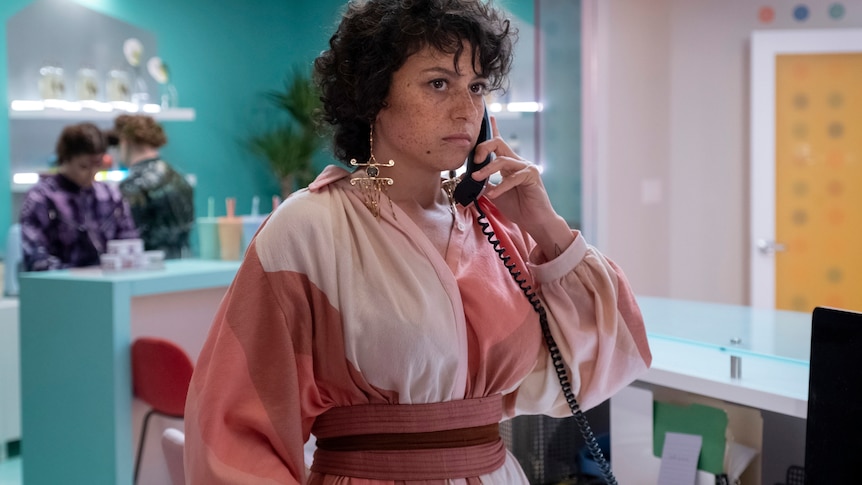 A young woman with black curly hair looks serious as she talks on the phone in a brighltly coloured room.