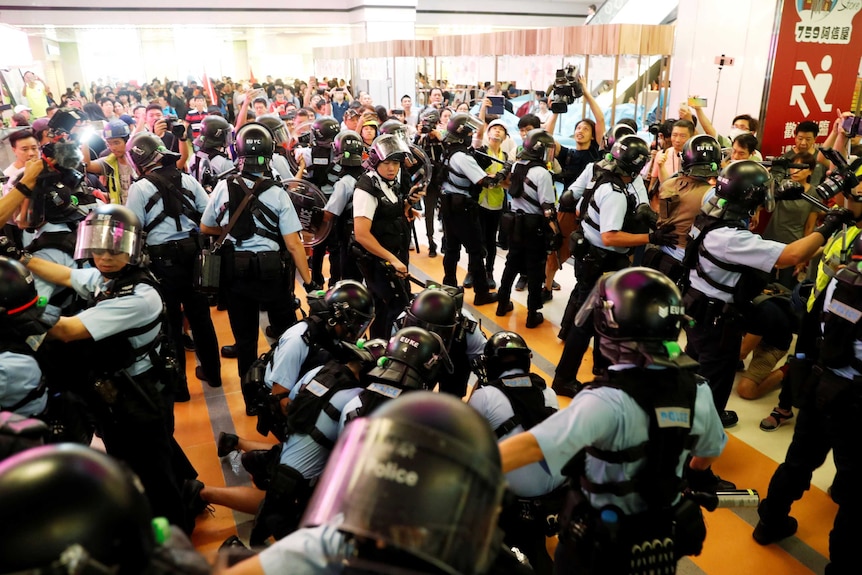 In a shopping centre, a vast crowd of police in riot gear push onlookers away as another group of them pin someone down.