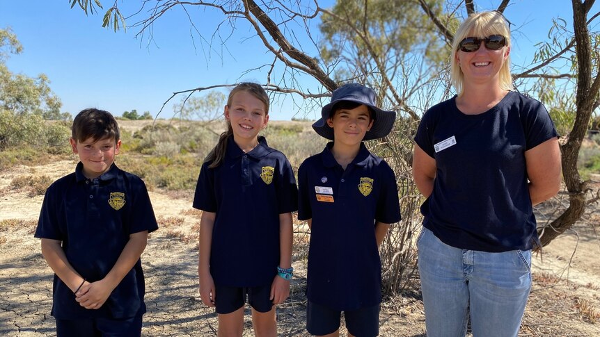 Three children and a woman stand next to each other smiling. There's a blue sky and dry trees and shrubs behind them.