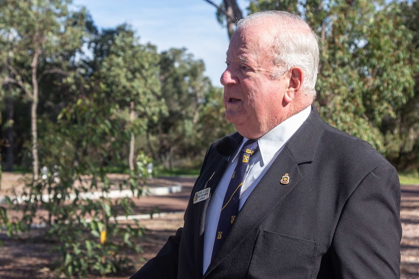 A middle-aged man with white hair, wearing a blazer standing in a park