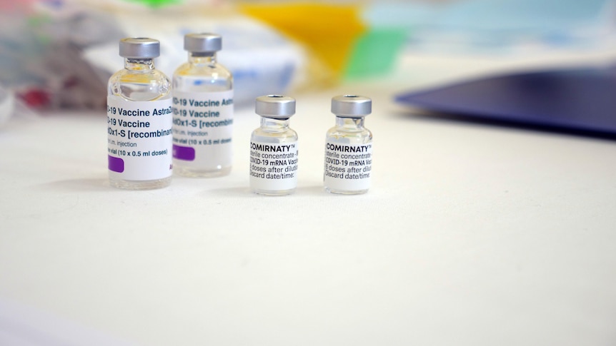 Vials of two different vaccines sit on a table. Medical supplies blurred in the background.