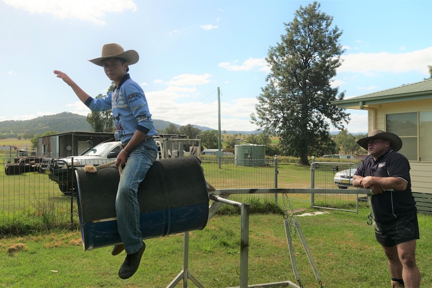 A boy wearing a cowboy hat sits atop a steel drum practicing bull riding as his father moves it. 