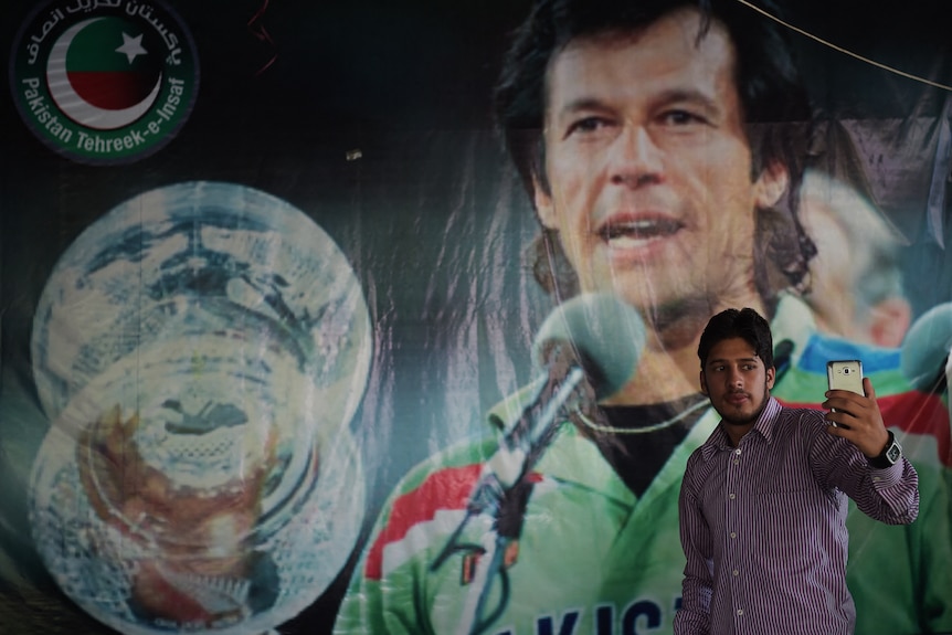 A man wearing a striped shirt holds up his phone in front of a sign of Imran Khan