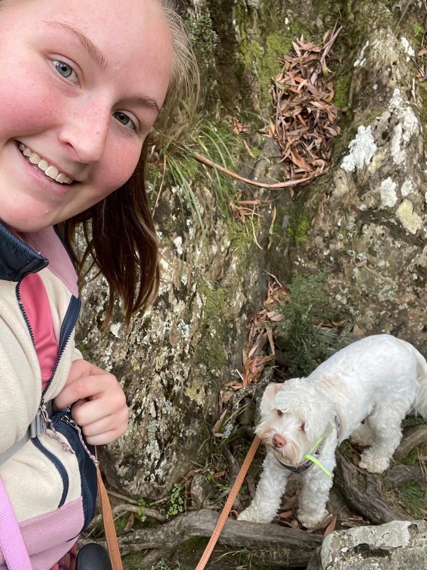 Woman smiling at camera beside her medium-sized white dog on a rocky trail.