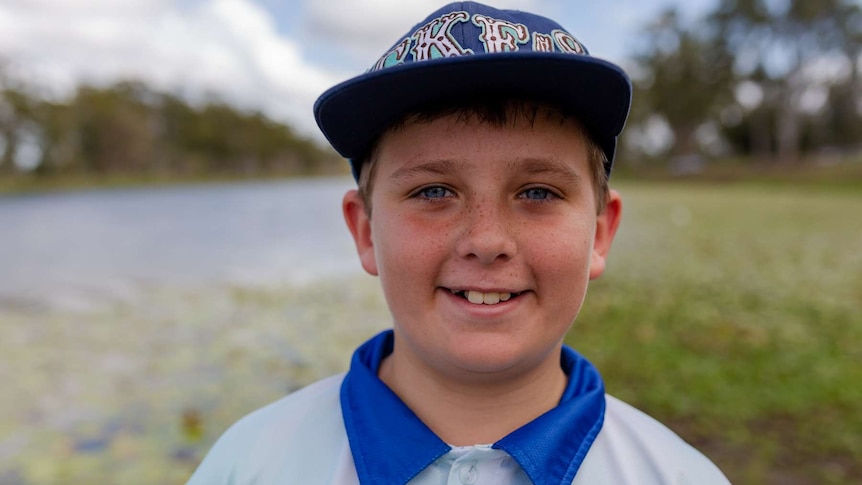 A young boy smiles at the camera, wearing a cap. Water and lilies in background blurred.