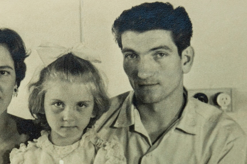 Black and white photo of young girl with stern expression holding one hand in her other, with parents smiling either side.