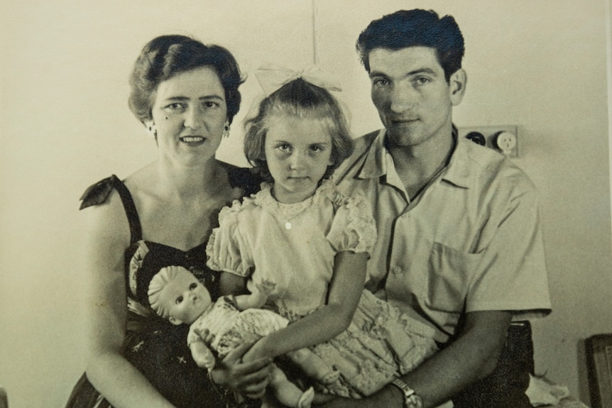 Black and white photo of young girl with stern expression holding one hand in her other, with parents smiling either side.
