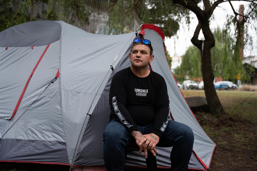 a-man-in-a-black-top-sits-in-front-of-grey-tend-in-a-park.