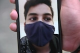 A young man's masked face appears on a video phone call held by a person's hand.