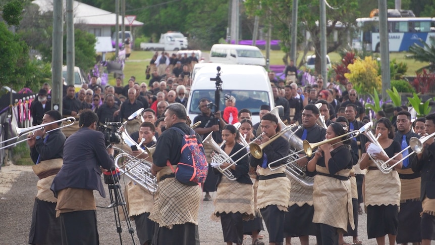 A funeral band is shown, playing instruments, and a crowd gathered behind them. 