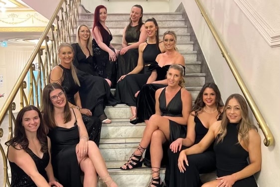 Eleven smiling young women, all wearing black dresses, sit on a flight of stairs.