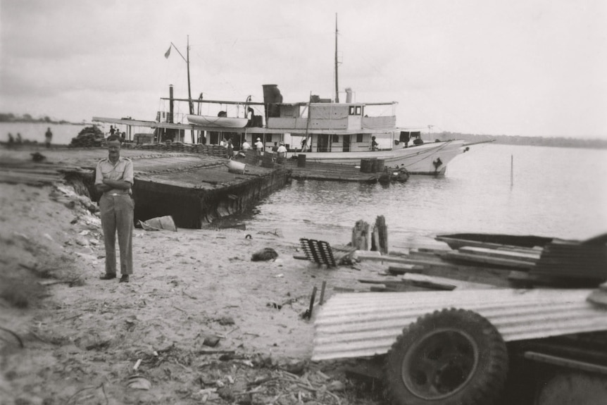 An old black and white photo of a man standing in a military uniform on a beach in front of a boat