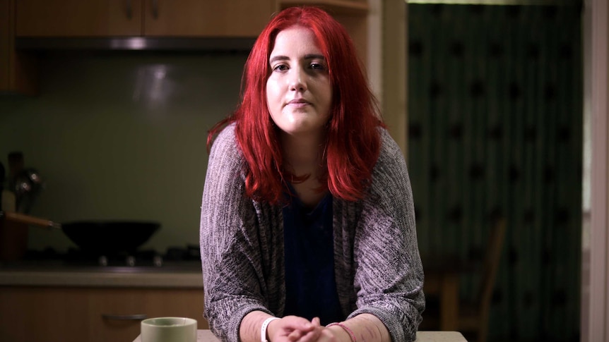 A young woman with bright red hair rests at a kitchen bench. Scars from her self-harming are just visible.