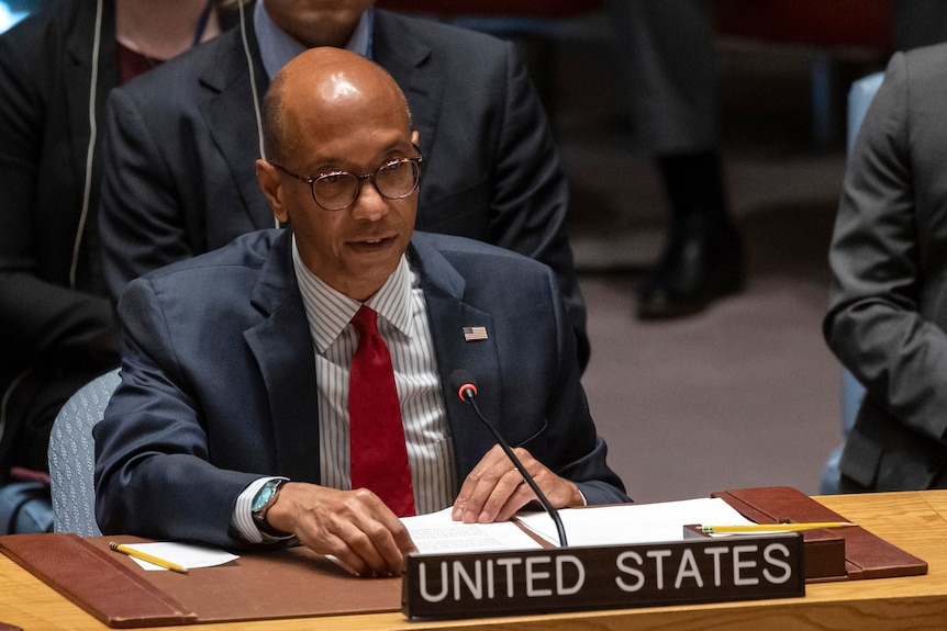 A man with bald head sits in front of sign reading "United States" at UN security council.