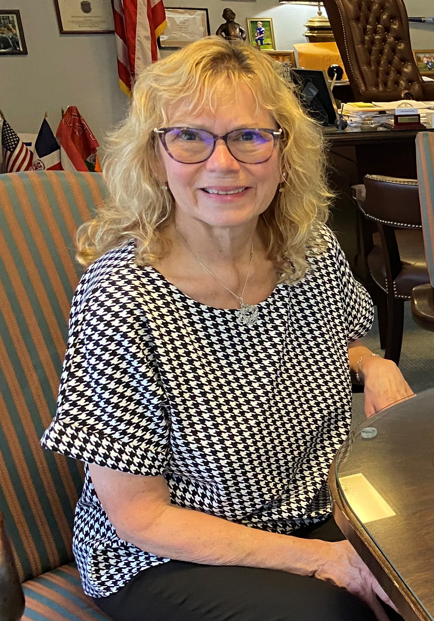 A woman in a black and white top and glasses is sitting and smiling.