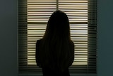 A woman stands before a window blind staring out.