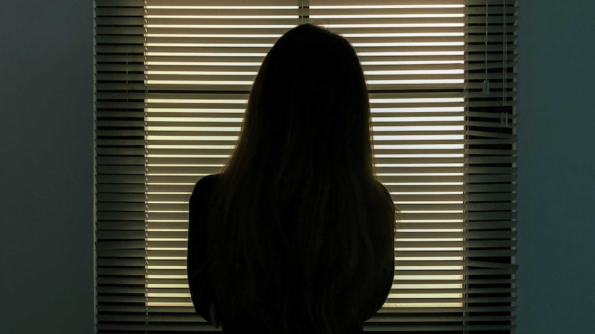 A woman stands before a window blind staring out.