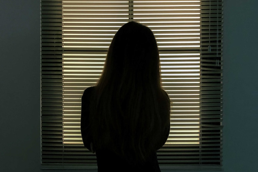 A woman stands before a window blind staring out.
