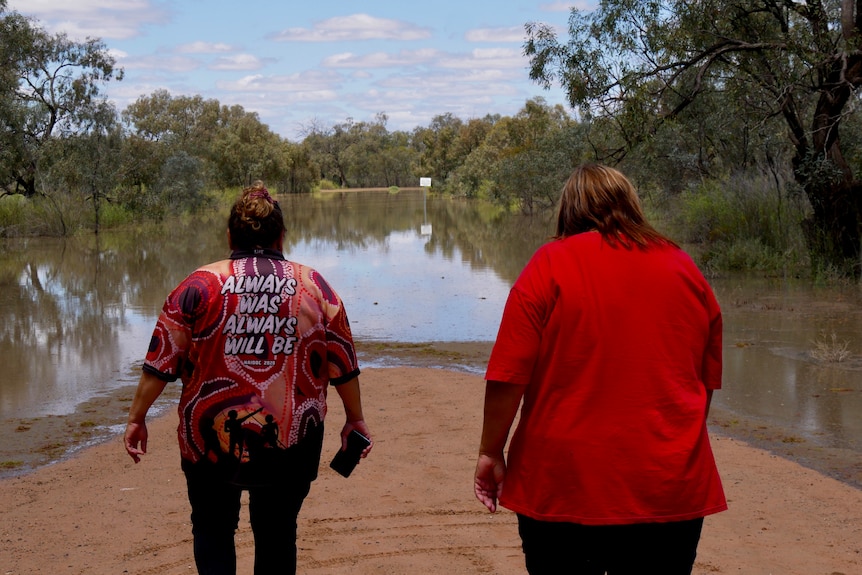 Two Indigenous women walking towards a road covered in water, their back towards the camera.