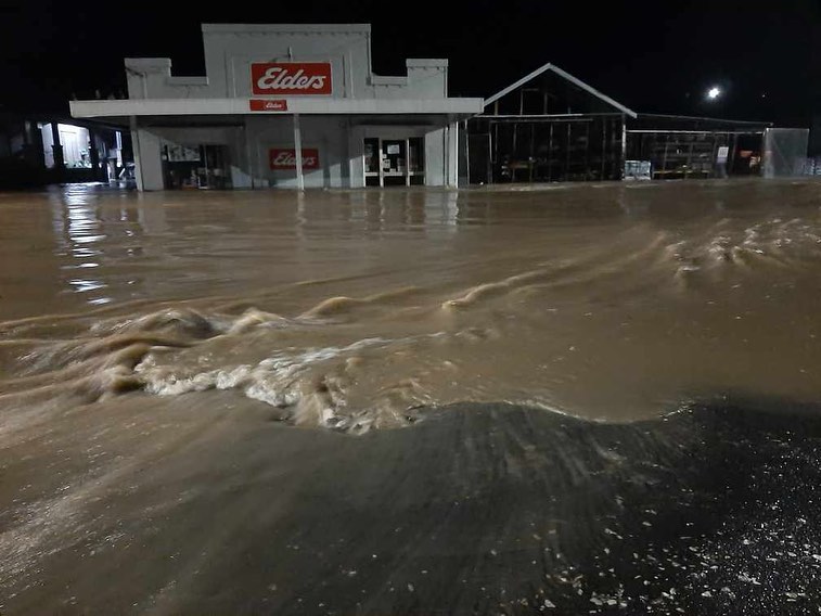 Deep brown flood water swirls around an Elders real estate store. The water level is halfway up the height of the shop's door.