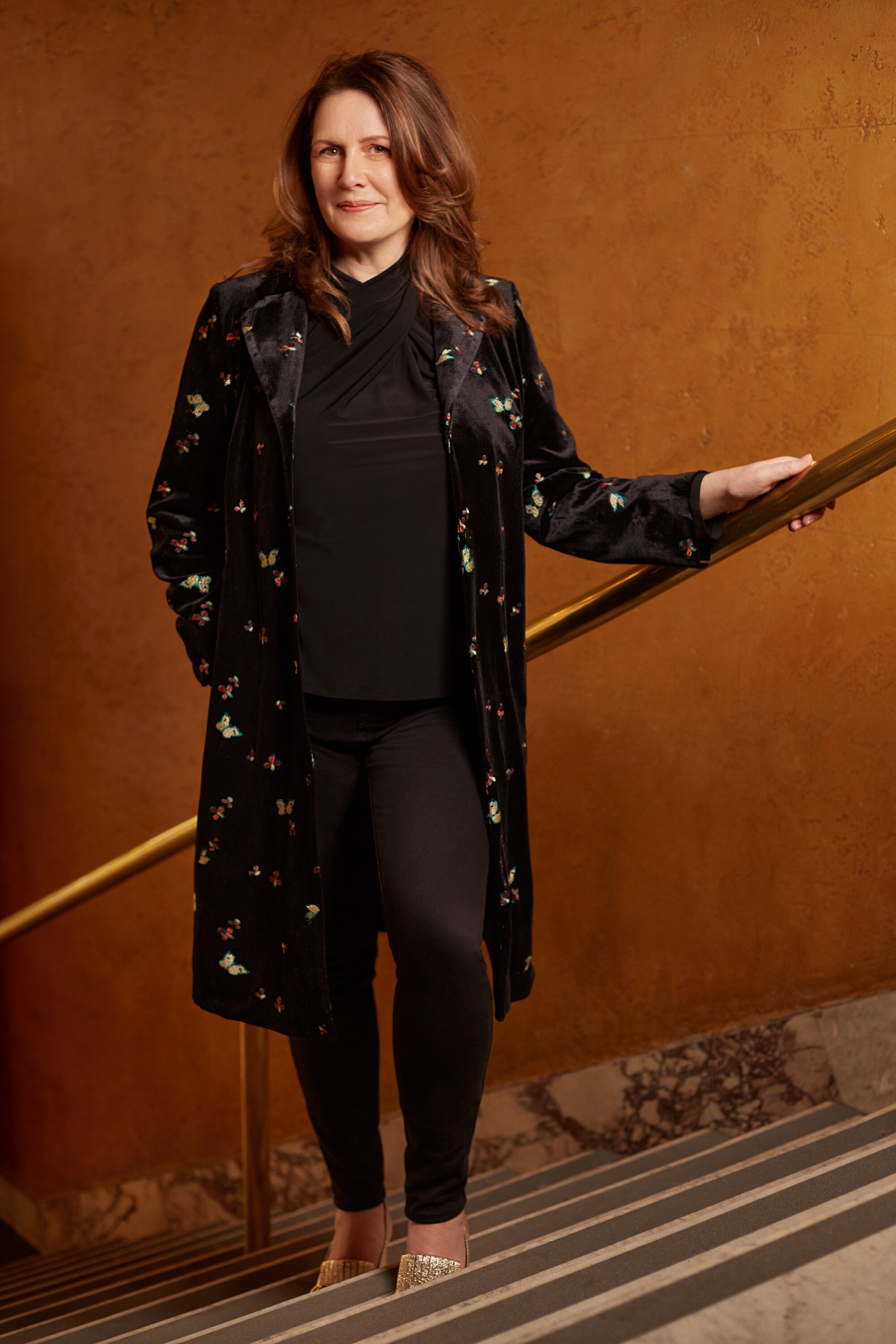 Carmen stands on the stairs with her hand on the railing. She has shoulder-length brown hair and is wearing black.