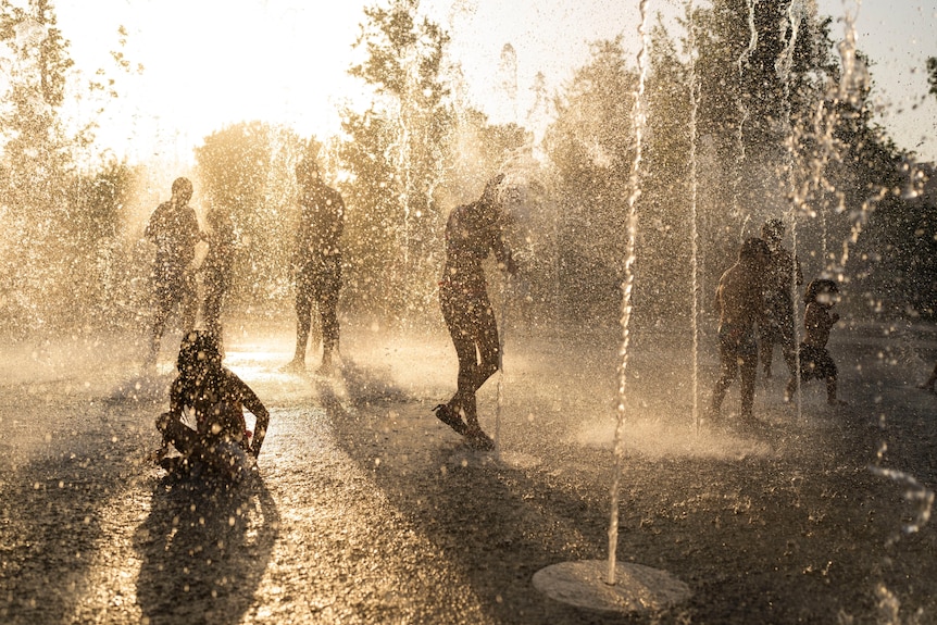 Silhouettes of children playing in water at a fountain in Athens during a heatwave with the sun behind them
