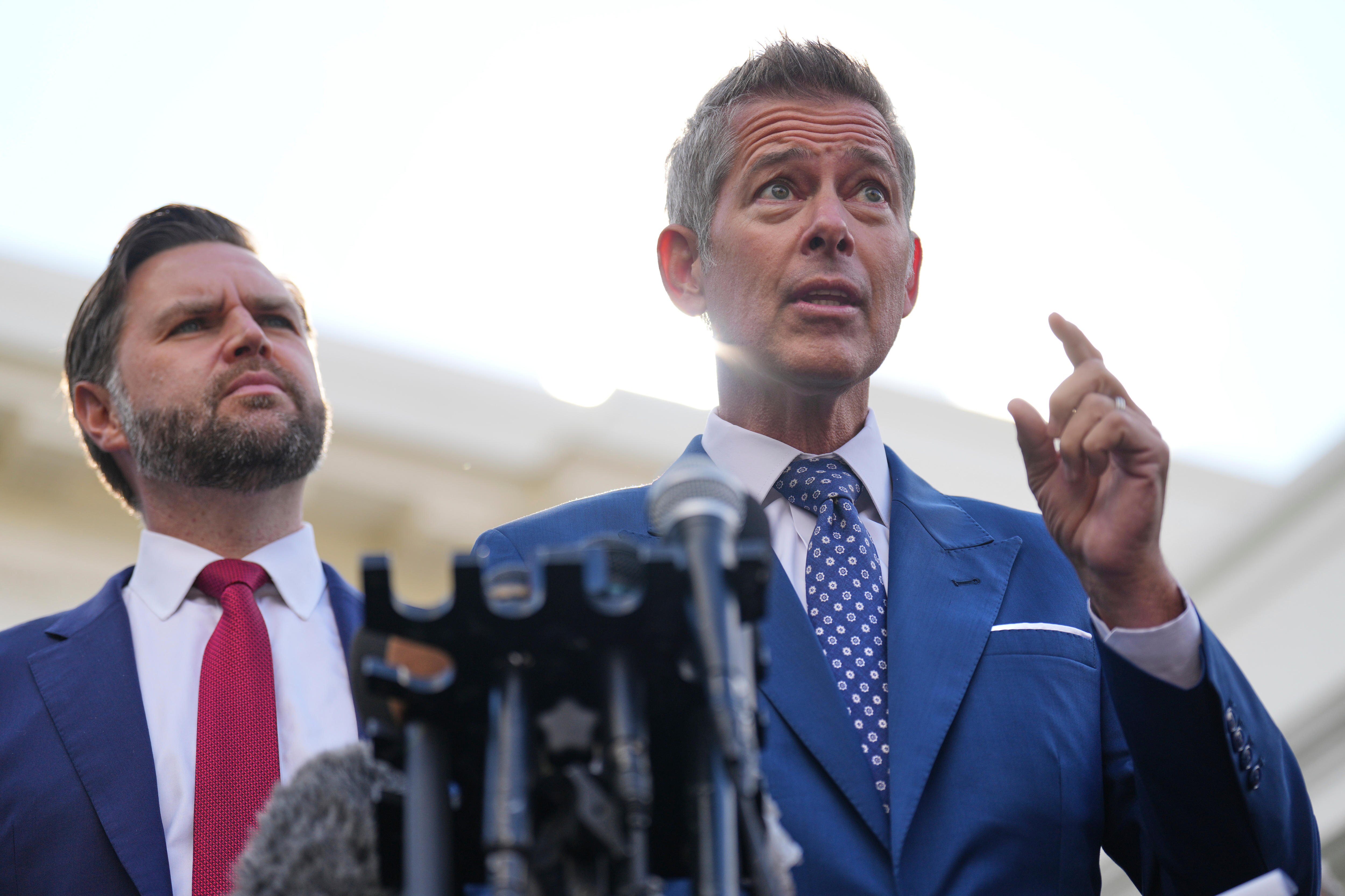 Sean Duffy in a blue suit and tie with his left arm and index finger raised, speaking next to a microphone and JD Vance.
