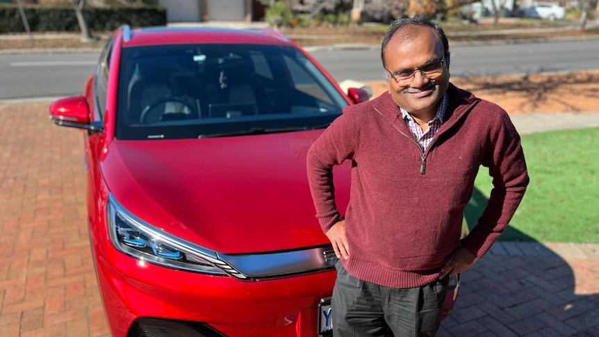 A man smiling and standing infront of a big red car