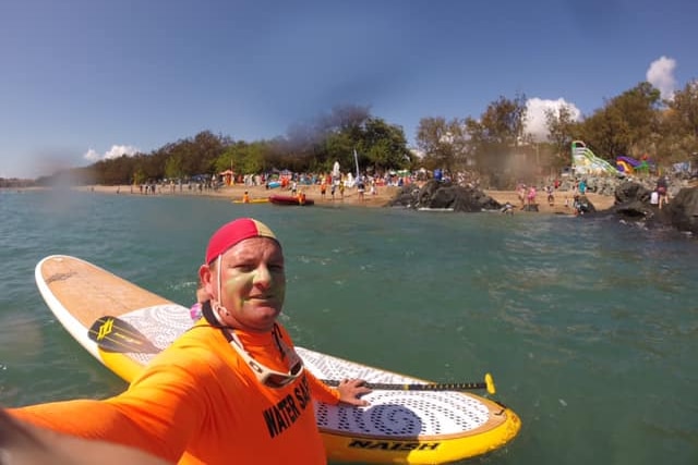 A smiling lifesaver treads water next to his board in the ocean.