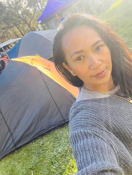 Analyn 'Logee' Osias smiles for a selfie with the sun softly shining on tents behind her.