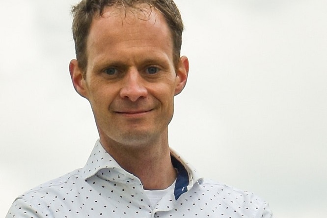 Een foto van een serieus uitziende man met terugwijkend bruin haar, witte overhemden met blauwe stippen, een wit T-shirt eronder.