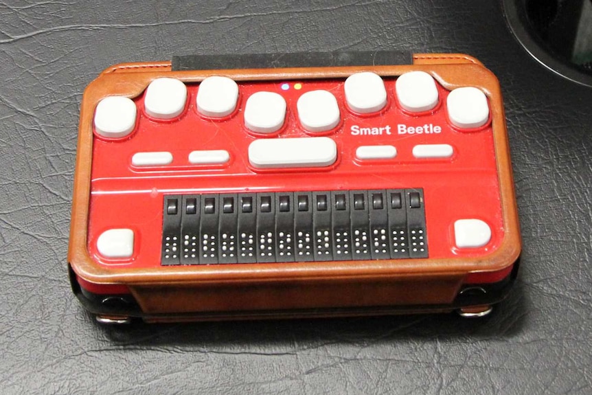 A red, rectangular device with 15 white buttons and 14 black cells which each contain braille.
