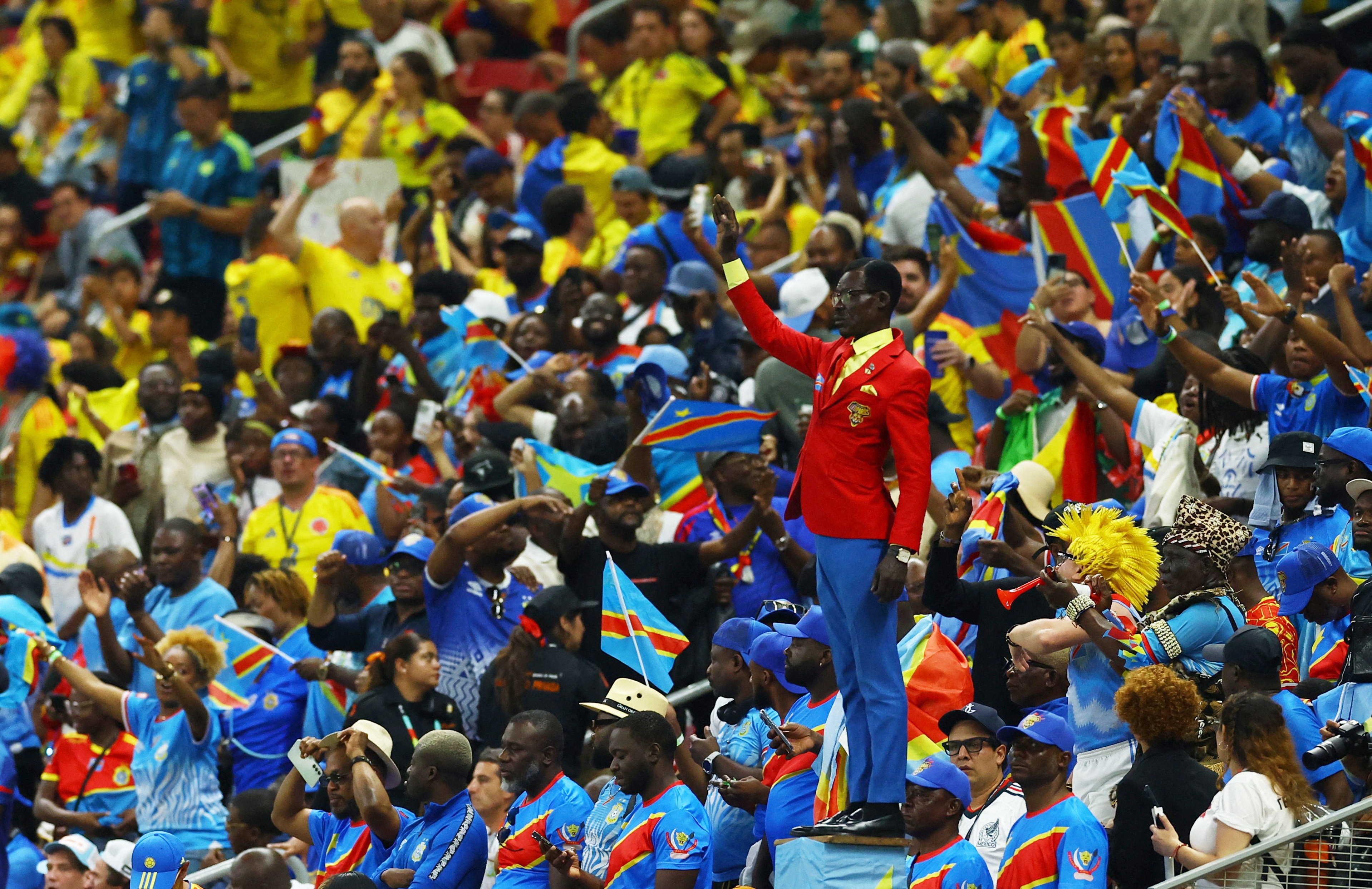 Michel Nkuka Mboladinga standing with one arm raised in a purple, blue and yellow suit