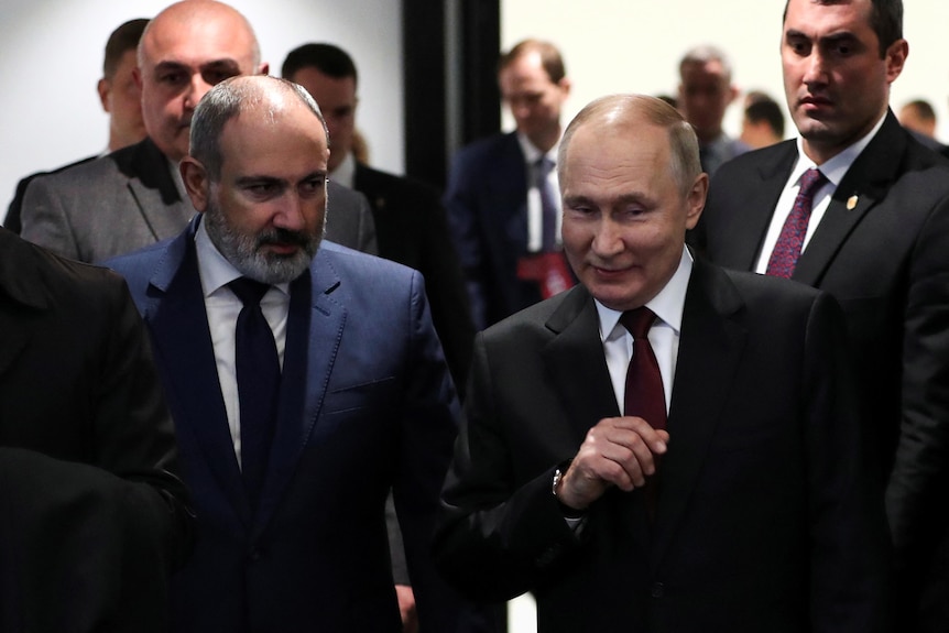 Vladimir Putin smirks as he walks beside another man in a suit, through a corridor