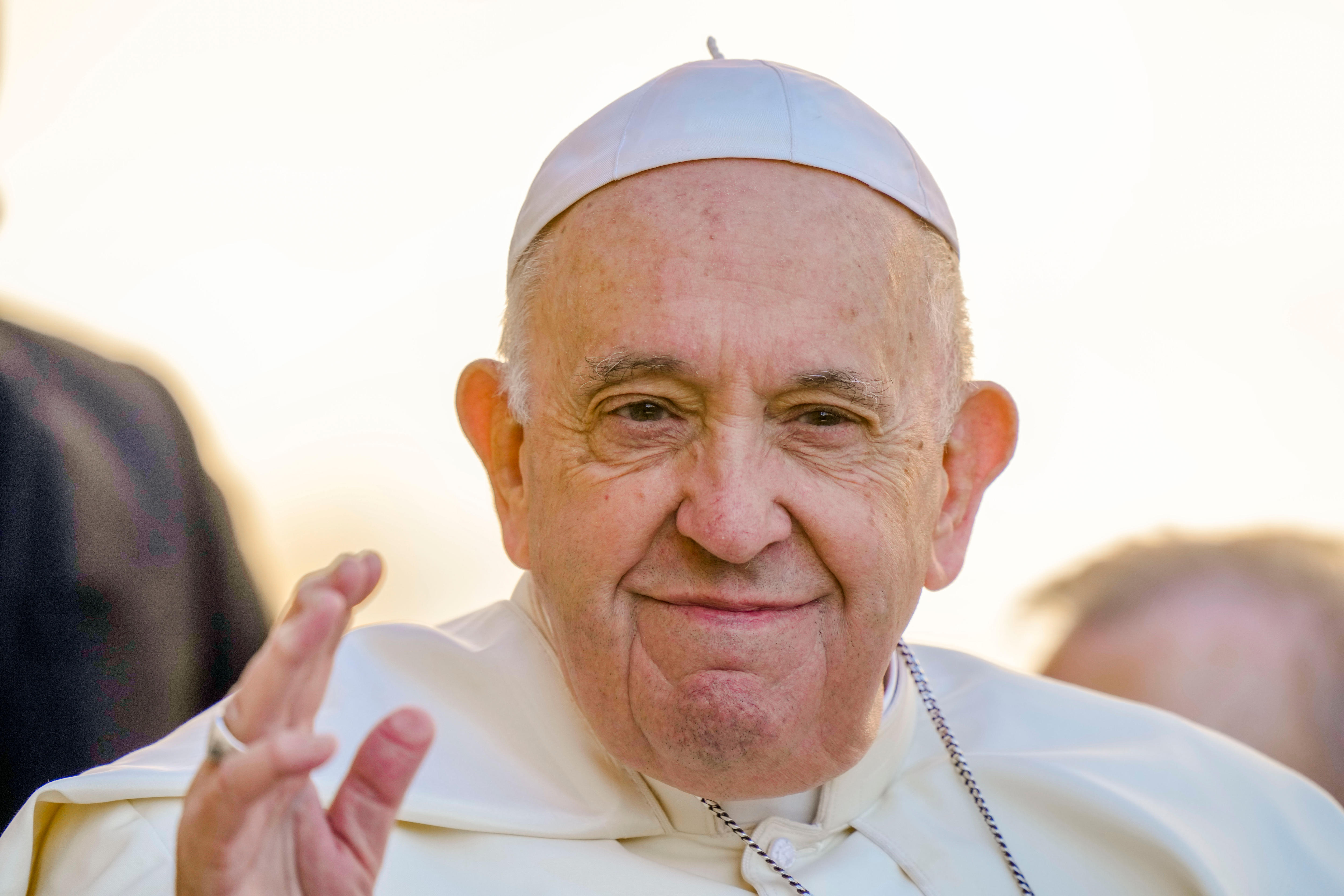 An elderly white man in a white gown and skull cap smiles as he waves in the direction of the camera.