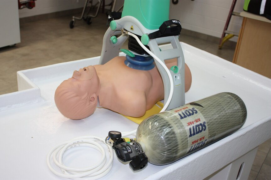 A plastic dummy lies on a medical table, next to a canister of oxygen and under the cardiac "thumper" machine