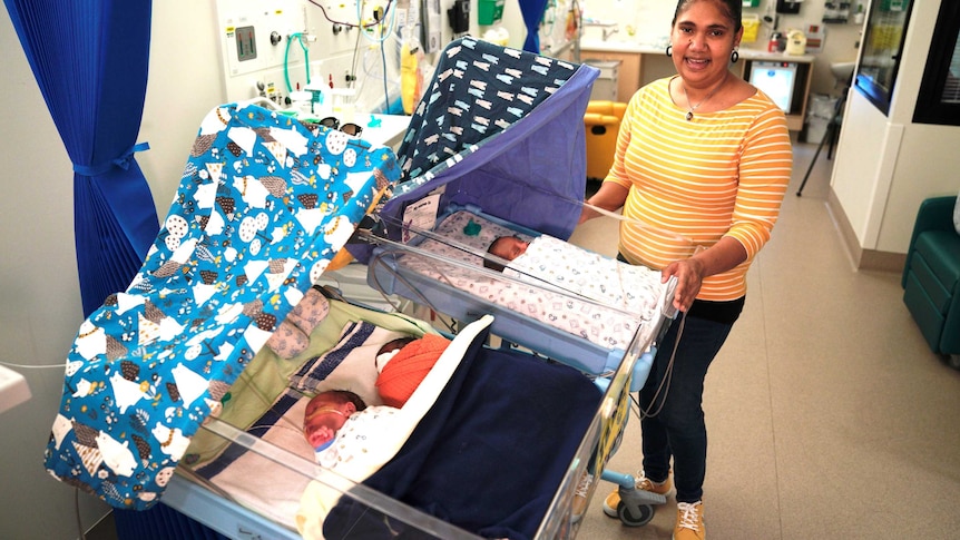 A smiling woman stands with three newborns sleeping in hospital cribs.