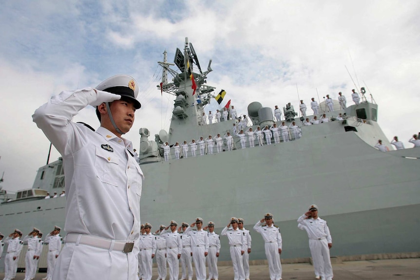 Sailor stands saluting in the foreground and group of saluting sailors stand before and on deck of a naval boat.